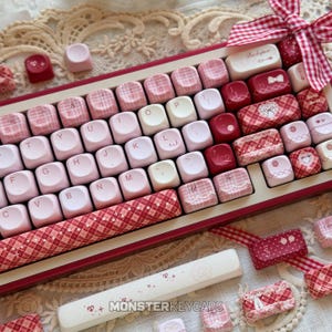 Puede incluir: Un teclado mecánico con un conjunto de teclas personalizadas en tonos rosa, blanco y rojo oscuro. Las teclas muestran una mezcla de colores sólidos, patrones de cuadros y pequeños iconos gráficos como lazos y corazones. Incluye una cinta de cuadros.