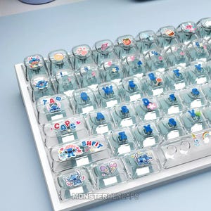 May include: A mechanical keyboard featuring transparent, light blue-tinted keycaps. Each keycap is decorated with small, colorful icons including cartoon characters, food items, and symbols. The keys display blue letters and the text SHIFT, CAPS, and TAB.