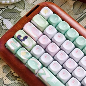 May include: A custom keyboard with pastel-colored keycaps. The keys are white, mint green, and pink, with printed letters and symbols. The keyboard sits in a wooden frame. Some keys have decorative patterns, including a squirrel and a hot air balloon.