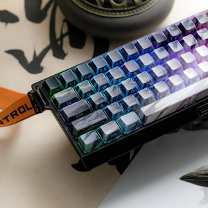May include: A mechanical keyboard featuring custom keycaps with a mountain landscape design and integrated RGB lighting. The keyboard has a black frame and an orange strap attached to the side with the text CONTROL visible on the fabric.