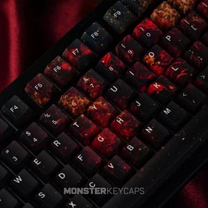 Puede incluir: Un teclado mecánico con teclas personalizadas que presentan un patrón jaspeado oscuro en tonos negro, rojo intenso y dorado. Las teclas muestran caracteres alfanuméricos y etiquetas blancas. El texto MONSTERKEYCAPS es visible en la parte inferior.