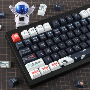 May include: Black mechanical keyboard with custom keycaps featuring space-themed designs and text. The keycaps are white, black, and red. An astronaut figurine is in the background. The keyboard has a variety of symbols and letters.