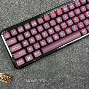 Puede incluir: Un teclado mecánico con teclas de color granate oscuro con letras y símbolos blancos. El teclado tiene un marco negro brillante y una retroiluminación roja que ilumina los espacios entre las teclas. El texto MONSTERKEYCAPS es visible en la parte inferior.