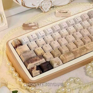 Peut inclure: Un clavier mécanique doté de touches personnalisées à l'esthétique vintage. Les touches présentent un mélange de tons crème, beige et marron, certaines affichant des portraits imprimés, du texte manuscrit et des motifs de partitions musicales.
