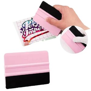 Puede incluir: La imagen muestra una espátula rosa y negra, con un mango de plástico rosa y una cuchilla de goma negra. La herramienta se utiliza para aplicar calcomanías de vinilo. Una mano sostiene la espátula y también se ve una calcomanía con el texto "Per Imper".