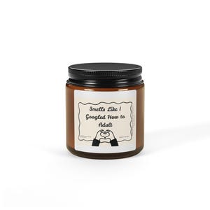 Puede incluir: Un tarro de vidrio marrón con tapa negra. La etiqueta dice "Smells Like I Googled How to Adult" con un gráfico de gesto de manos en forma de corazón. El tarro es probablemente una vela perfumada.