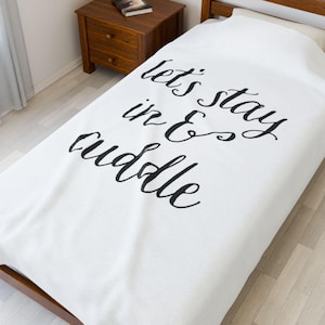 Puede incluir: Manta blanca con la frase "let's stay in & cuddle" en letras negras. La manta está sobre una estructura de cama de madera. Una pequeña mesita de noche de madera con un libro es visible en el fondo.