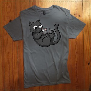 May include: A gray t-shirt featuring a cartoon cat and mouse graphic. The cat is dark gray with large eyes, holding a small gray mouse. The design is centered on the shirt, which is laid flat on a wooden surface.