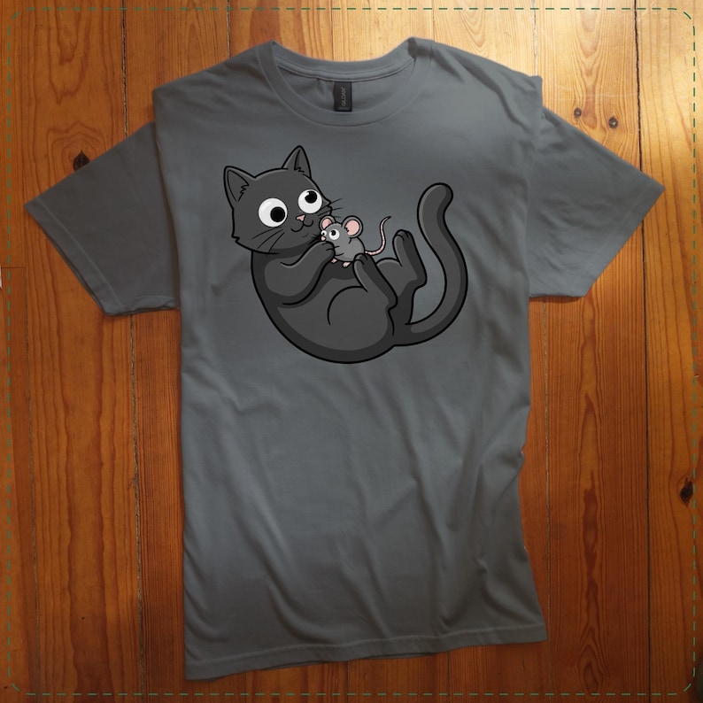 May include: A gray t-shirt featuring a cartoon cat and mouse graphic. The cat is dark gray with large eyes, holding a small gray mouse. The design is centered on the shirt, which is laid flat on a wooden surface.
