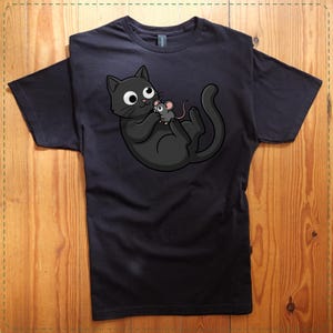May include: Black t-shirt featuring a cartoon cat holding a mouse. The cat is black with large white eyes and a playful expression. The mouse is gray with pink ears and a long tail. The design is centered on the shirt.