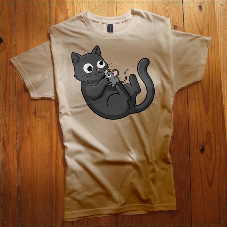 May include: Tan t-shirt featuring a cartoon illustration of a black cat holding a gray mouse. The cat has large, round eyes and a playful expression. The mouse is small and gray. The design is centered on the shirt.