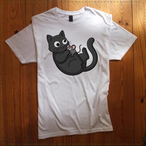 May include: White t-shirt featuring a cartoon illustration of a black cat holding a gray mouse. The cat has large eyes and a smiling expression. The shirt is made of a soft material and has a crew neck.