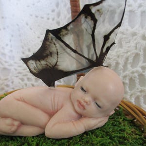 May include: A figurine of a sleeping baby with a butterfly wing attached to its back, resting on green moss inside a woven basket. The butterfly wing is black and white. The baby has closed eyes and is lying on its side.