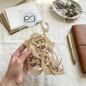 May include: A wooden art piece with an abstract design, held in front of a clear acrylic sheet. The artwork features flowing lines and curves. The logo "p3 design" is visible on the acrylic. The background includes stationery and a leather notebook.