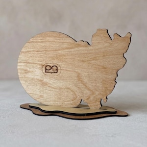 May include: A light brown wooden cutout shaped like a snowman, featuring a circular head and a jagged, irregular body. The piece is mounted on a flat, dark-edged wooden base. A small logo with the text "PG design" is engraved on the front.