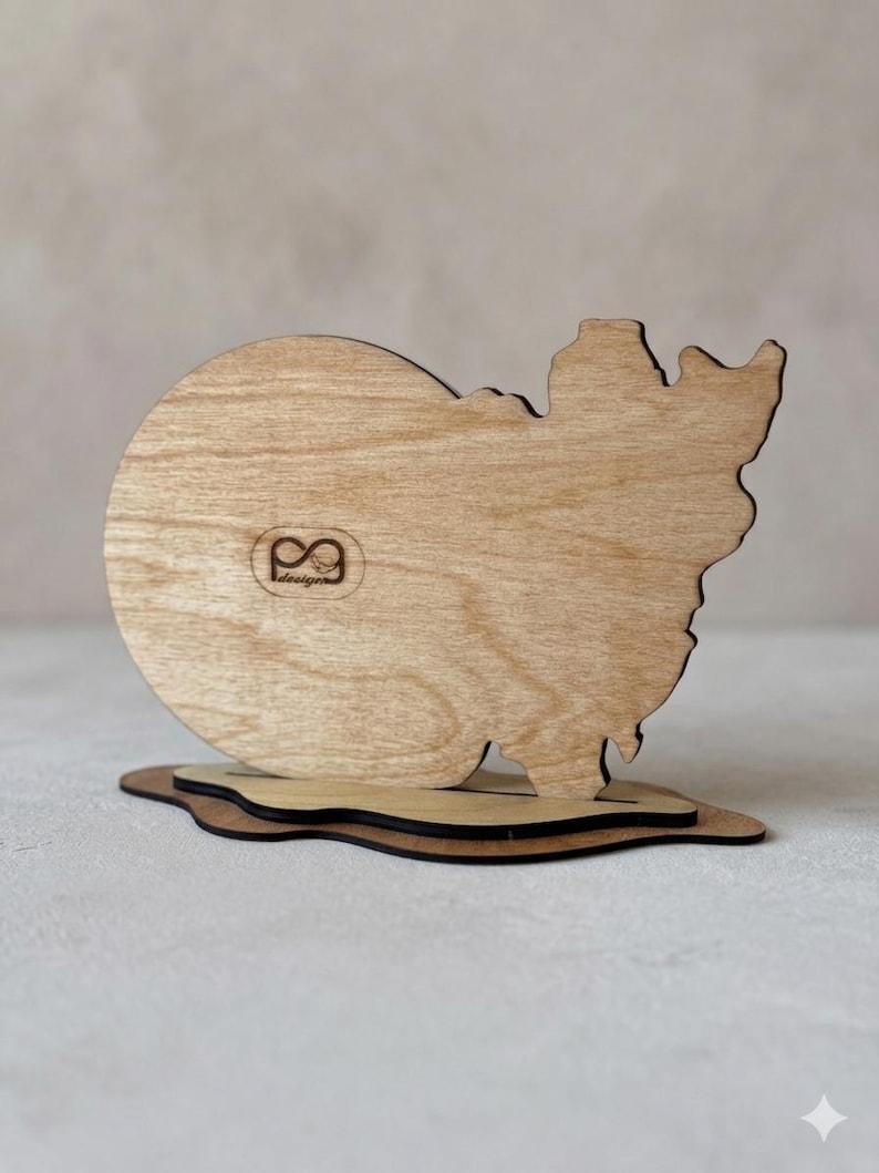 May include: A light brown wooden cutout shaped like a snowman, featuring a circular head and a jagged, irregular body. The piece is mounted on a flat, dark-edged wooden base. A small logo with the text "PG design" is engraved on the front.