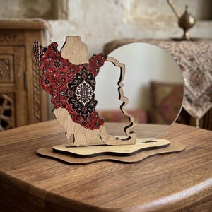 May include: A decorative tabletop mirror featuring a wooden frame shaped like the map of Iran. The left side of the wooden frame is decorated with a detailed red, black, and white floral pattern, while the right side holds a circular mirror.