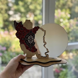 May include: A decorative wooden stand featuring a cutout map of Iran on the left side, decorated with a red, black, and white intricate floral pattern. A circular mirror is attached to the right side, all mounted on a layered, wavy wooden base.