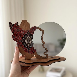 Op de afbeelding: Een decoratieve tafelspiegel met een ronde glazen ruit bevestigd aan een houten frame. Het frame heeft aan één kant een onregelmatige, organische vorm, versierd met een gedetailleerd rood, zwart en wit bloemenpatroon op het hout.
