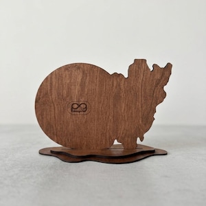 May include: A dark brown wooden display stand featuring a circular left side and an irregular, jagged right side. The piece is mounted on a layered, wavy wooden base. A small logo with the text "pg designs" is engraved on the circular section.