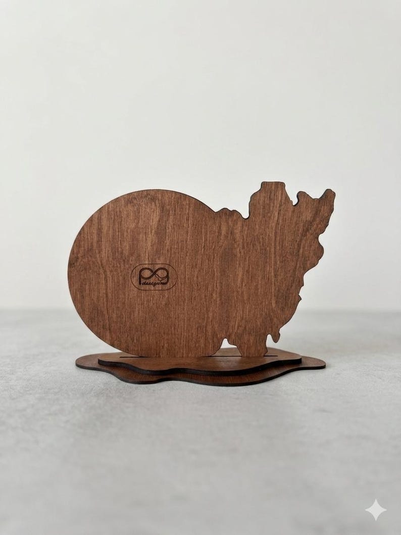 May include: A dark brown wooden display stand featuring a circular left side and an irregular, jagged right side. The piece is mounted on a layered, wavy wooden base. A small logo with the text "pg designs" is engraved on the circular section.