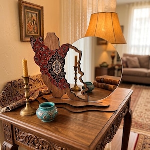 May include: A circular mirror with a wooden frame shaped like the map of Iran, featuring a red, black, and white intricate floral pattern on the left side. The mirror is mounted on a wooden base and sits on a carved wooden table.