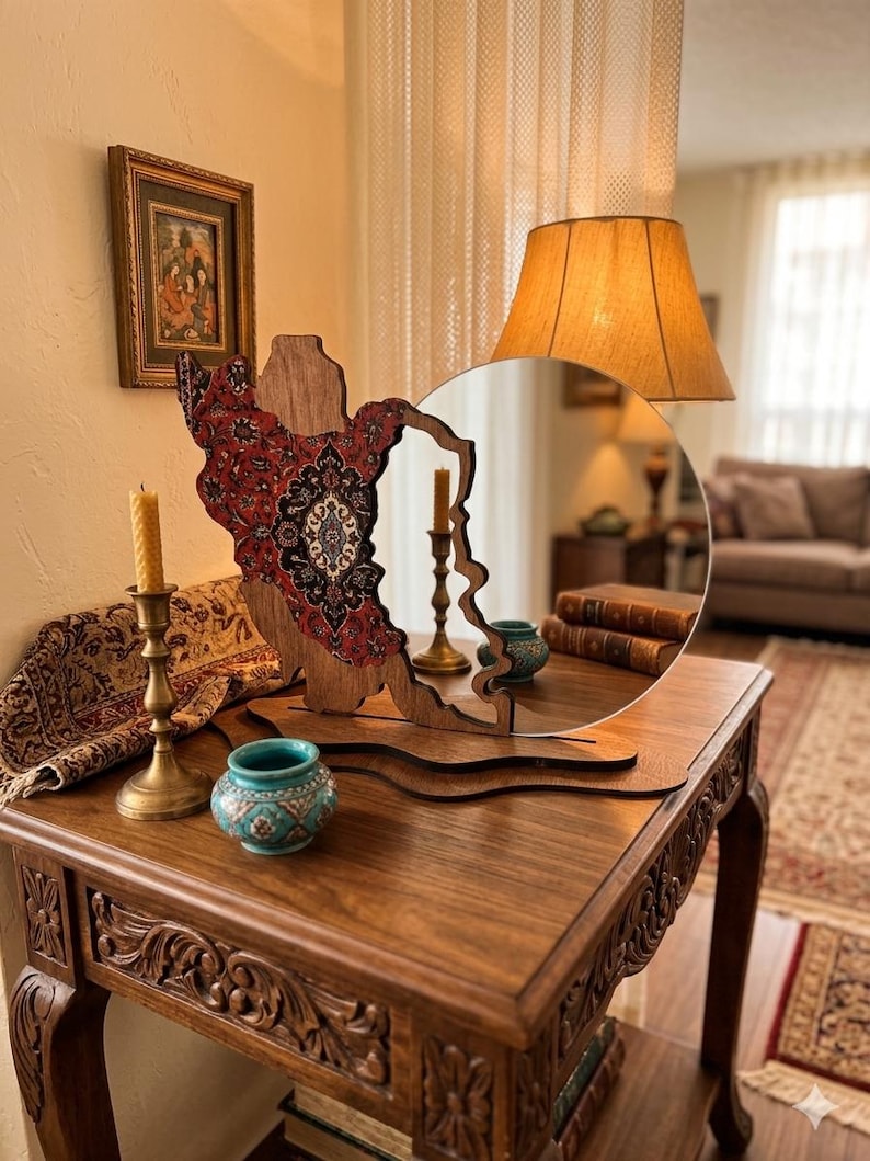 May include: A circular mirror with a wooden frame shaped like the map of Iran, featuring a red, black, and white intricate floral pattern on the left side. The mirror is mounted on a wooden base and sits on a carved wooden table.
