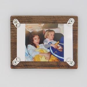 Puede incluir: Un marco de fotos rústico de madera con una fotografía de dos niños. El marco es de madera marrón oscuro y envejecida, asegurado con pestañas de metal. La fotografía está centrada dentro del marco.