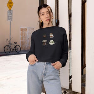 Puede incluir: Sudadera corta negra con cuatro diseños de gatos con temática de café. Las ilustraciones incluyen gatos en una taza de café, un café con leche y una taza de té. La sudadera tiene mangas largas y cuello redondo.