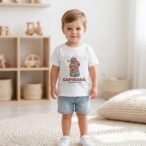 May include: A white t-shirt featuring a cartoon capybara design with the text "CAPYBARA ARGENTINA". The shirt is worn by a young child, paired with denim shorts and white sneakers. The background includes a light-colored rug and wooden shelves.