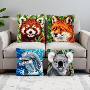 Op de afbeelding: Vier vierkante kussens met dierenontwerpen. De kussens hebben een rode panda, een knipogende vos, een dolfijn en een koala. Elk kussen heeft een groene achtergrond met een getextureerd, handgemaakt uiterlijk.