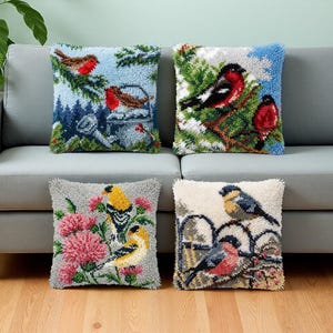 Könnte beinhalten: Vier quadratische Kissen mit texturierten Punch-Needle-Motiven verschiedener Vögel. Oben links Rotkehlchen in einer Winterszene, oben rechts Gimpel auf Zweigen, unten links Stieglitze mit rosa Blumen und unten rechts Vögel auf einem Zaun.