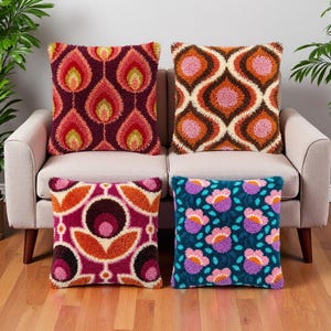 May include: Four square throw pillows with textured punch needle embroidery. The top left features red and yellow flame shapes on a dark purple background. The top right has orange and pink geometric patterns. The bottom left shows pink and orange floral motifs on a magenta base. The bottom right displays pink and purple flowers on a dark teal background.