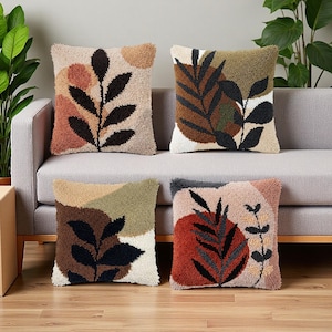 Könnte beinhalten: Vier quadratische Kissen mit strukturierter Punch-Needle-Stickerei und abstrakten botanischen Blattmustern. Die Designs enthalten erdige Farbpaletten mit Tönen in Terrakotta, Olivgrün, Creme, Braun und Schwarz auf einer getufteten Oberfläche.