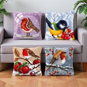 Op de afbeelding: Vier vierkante kussens met getextureerde punch needle ontwerpen van vogels in winterse taferelen. De ontwerpen tonen roodborstjes en een pimpelmees op besneeuwde takken met rode bessen en appels, uitgevoerd in levendige garenkleuren.