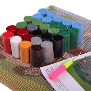 May include: A latch hook craft kit featuring a grid canvas, a printed instruction sheet, and a metal hook tool with a pink and yellow handle. The kit includes numerous pre-cut yarn bundles in various colors, including red, white, orange, gray, black, blue, and green, arranged in a cluster.