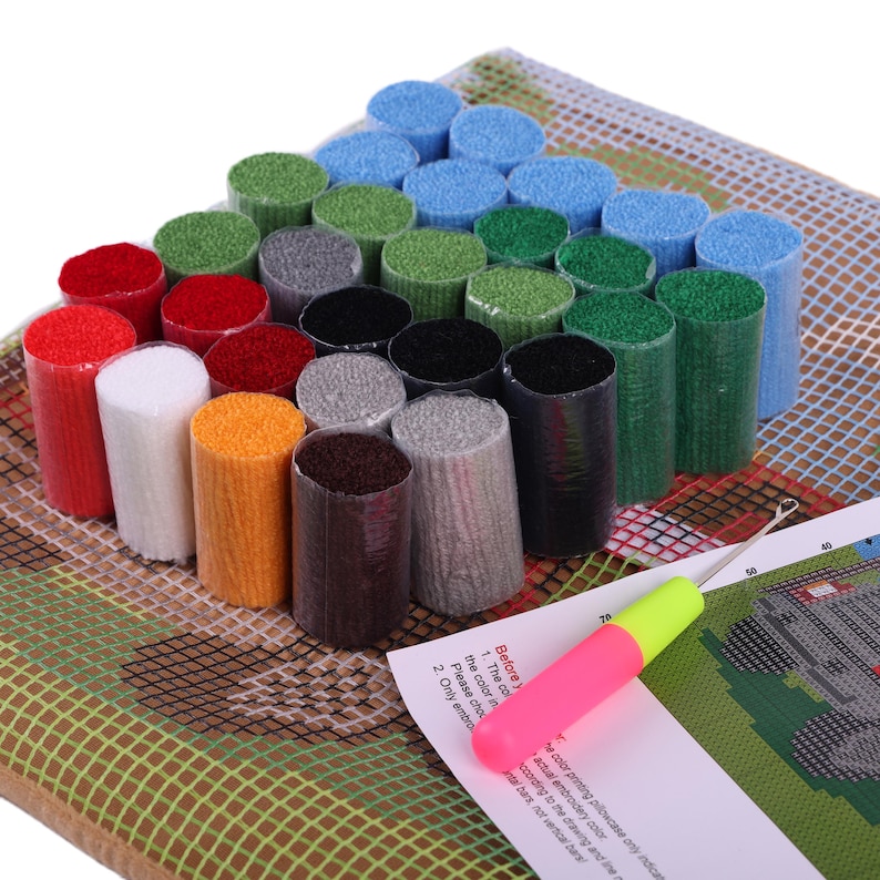 May include: A latch hook craft kit featuring a grid canvas, a printed instruction sheet, and a metal hook tool with a pink and yellow handle. The kit includes numerous pre-cut yarn bundles in various colors, including red, white, orange, gray, black, blue, and green, arranged in a cluster.