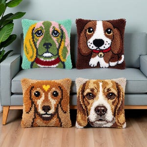 Peut inclure: Quatre coussins carrés présentant des motifs en punch needle texturé représentant des visages de chiens. Le coussin en haut à gauche montre un chien vert et jaune, celui en haut à droite un chien marron et blanc, en bas à gauche un chien fauve et à droite un beagle.