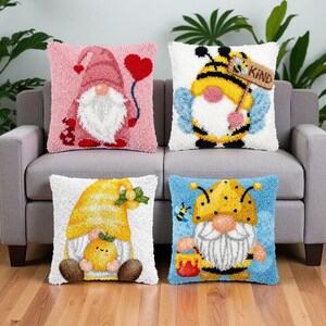 May include: Four square throw pillows featuring textured punch needle designs. One is pink with a gnome holding a red heart. One is white with a bee-themed gnome holding a sign that says KIND. One is white with a yellow gnome holding a small yellow creature. One is blue with a bee-themed gnome holding a honey pot.