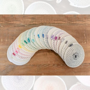 Op de afbeelding: Een set van 18 ronde, geweven katoenen onderzetters in een regenboog van kleuren. De onderzetters zijn in een halve cirkel gestapeld, met de zwart-witte onderzetter onderaan.