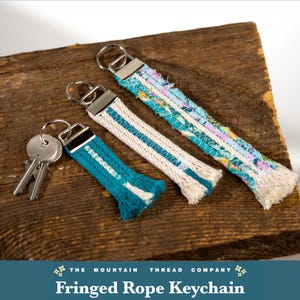 May include: Three keychains with silver-toned metal rings and rectangular clasps. The keychains feature frayed rope and fabric strips in teal, white, and floral patterns. The smallest keychain is attached to two silver-toned metal keys.