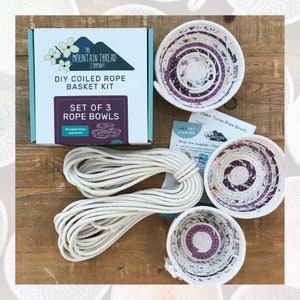 May include: A DIY coiled rope basket kit from The Mountain Thread Company. The kit includes three pre-made rope bowls in a purple and white pattern, a roll of white rope, and instructions. The kit is on a wooden surface.