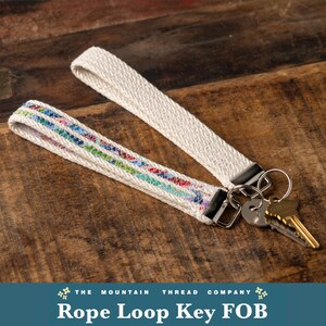 May include: Two wristlet key fobs made of off-white woven rope. One fob features a colorful, multi-patterned stripe design, while the other is solid off-white. Both include silver-toned metal hardware and a split ring holding two brass keys.