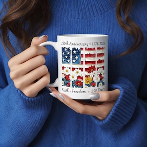 May include: White ceramic mug featuring a patriotic design with an American flag motif, daisies, and text that reads "250th Anniversary 1776-2026 Faith Freedom 250 Years". The mug is held by a person wearing a blue sweater.