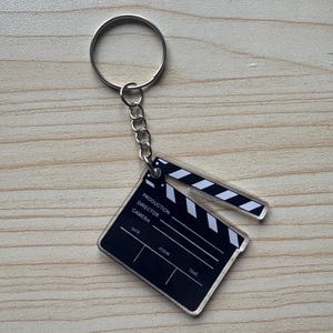 May include: A black and white film clapperboard keychain. The keychain features a rectangular clapperboard with the words "PRODUCTION, DIRECTOR, CAMERA" and fields for "DATE, SCENE, TAKE". The clapperboard is attached to a silver chain and keyring.