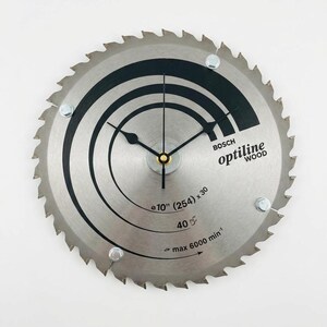 Peut inclure: Une lame de scie circulaire transformée en horloge. La lame argentée présente des lignes noires incurvées et le texte "BOSCH optiline WOOD". Les aiguilles de l'horloge sont noires, et le diamètre est de 25,4 cm.