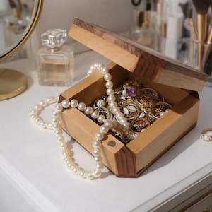 May include: A wooden jewellery box with an open lid, filled with various rings and necklaces. A pearl necklace drapes over the side. A gold ring with a pearl sits nearby. The box is on a white surface.