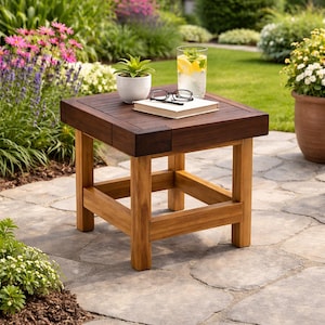 May include: A square wooden side table with a dark brown top and light brown legs. A small potted plant, a book, and a glass of water with lemon slices rest on the table. The table is outdoors on a stone patio.