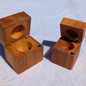 May include: Two small, open wooden boxes with hinged lids. The boxes are square with a circular indentation in the base and lid. The wood has a warm, reddish-brown tone. Small magnets secure the lids.