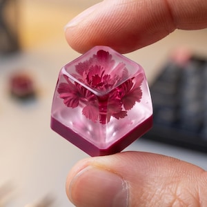 May include: A clear, square resin keycap with a deep red flower encased inside. The keycap has a dark red base and is held between fingers. The background is blurred, showing a desk with office supplies.